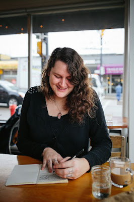 A dark haired woman in a black shirt writing in a notebook while sitting at a table in a restaurant.